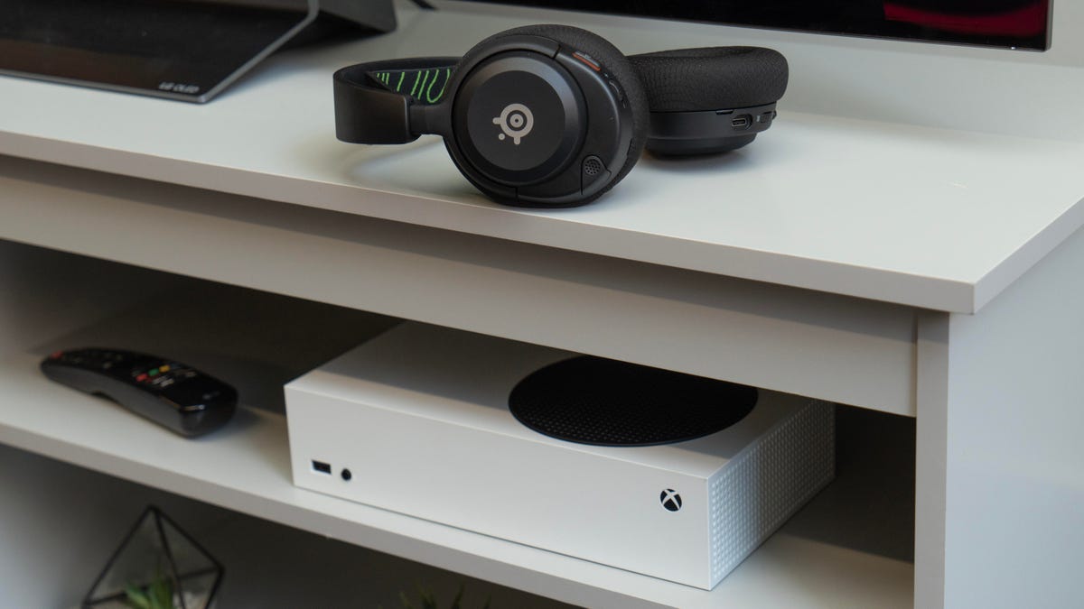 The Arctis Nova 5X rests on a gray media center, the right earcup facing you and the left earcup turned face up, with a white Xbox Series S sitting on the shelf below it.