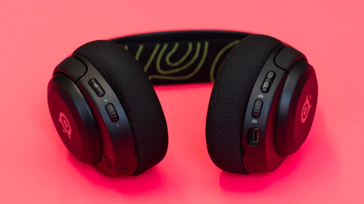 An Arctis Nova 5X headset set against a hot pink background with the headset controls facing you, with a microphone tucked into the earpiece.  Controls are volume, mic mute, power, link game/chat balance.