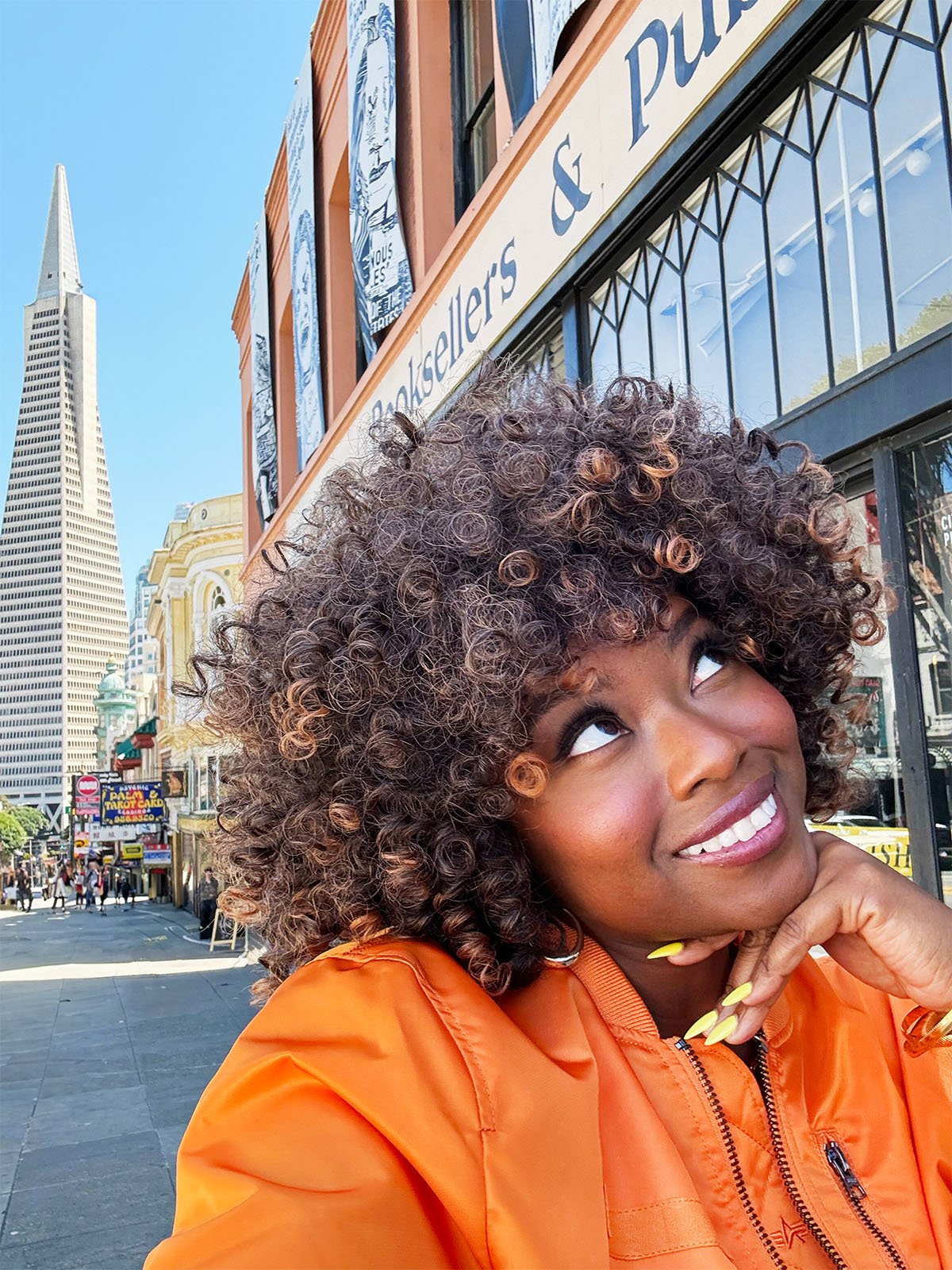 A woman with curly hair and an orange jacket smiles as she looks up.  She is standing on a city street with a tall, pointed skyscraper in the background.  You can also see the facade of a bookstore.