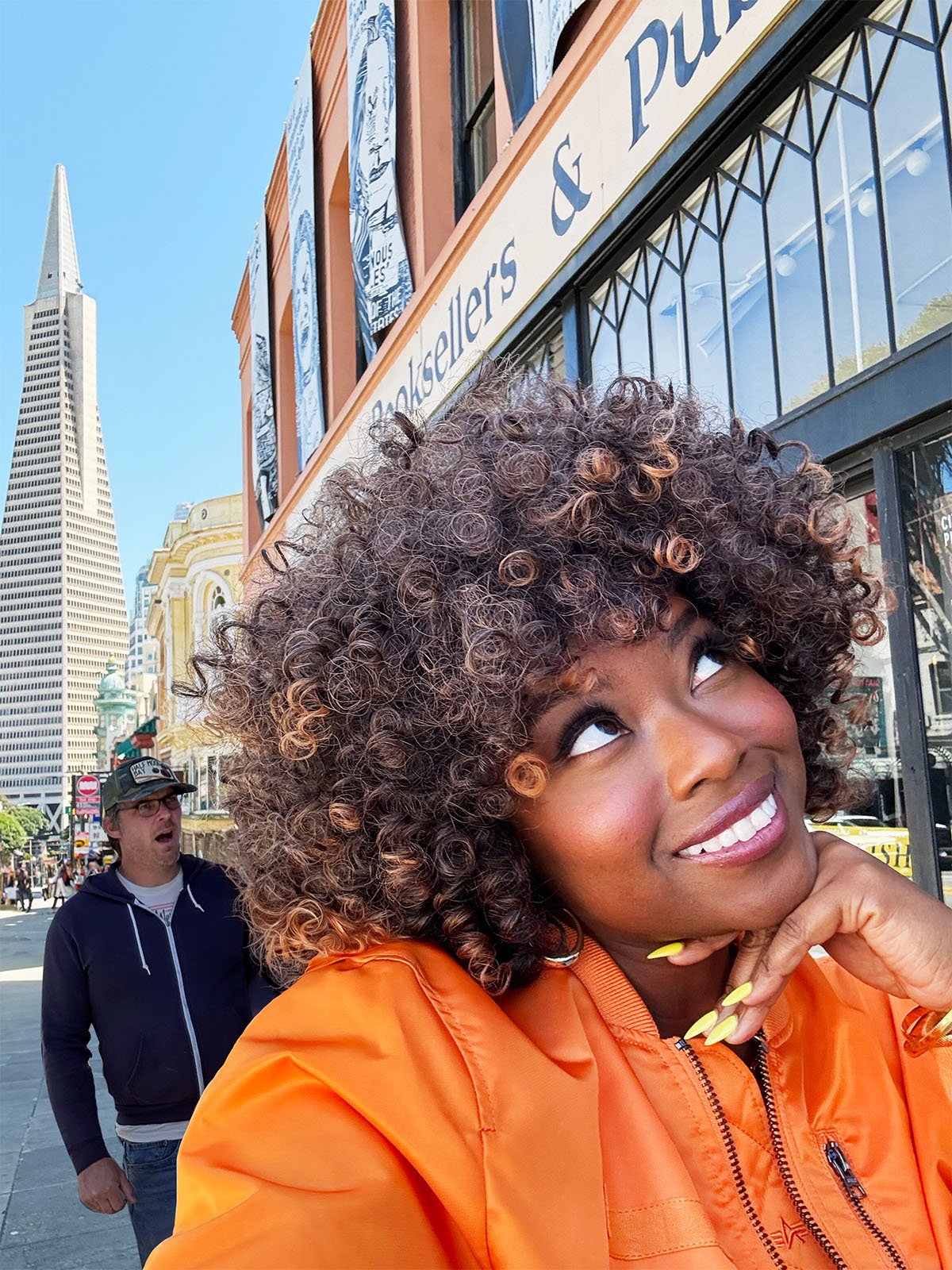 A woman with curly hair and an orange jacket smiles as she looks up, standing in front of a building with a sign that reads "Bookstores and publishers." A hooded man appears surprised in the background.  Behind them is the Trans-American Pyramid.