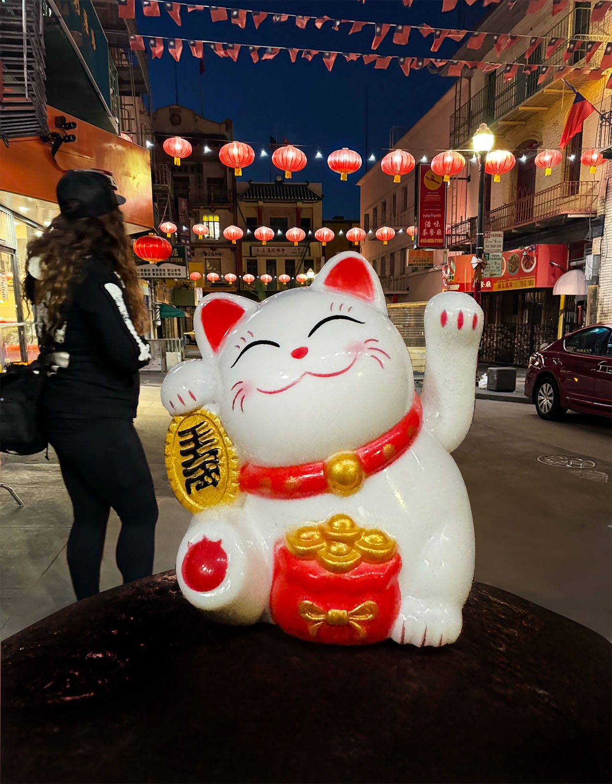 A large Maneki-neko (baiting cat) statue with raised paw and red collar stands prominently on a street lit by lanterns.  In the background, a man dressed in black clothing and a hat walks away, red lanterns hanging above him, illuminating the evening scene.