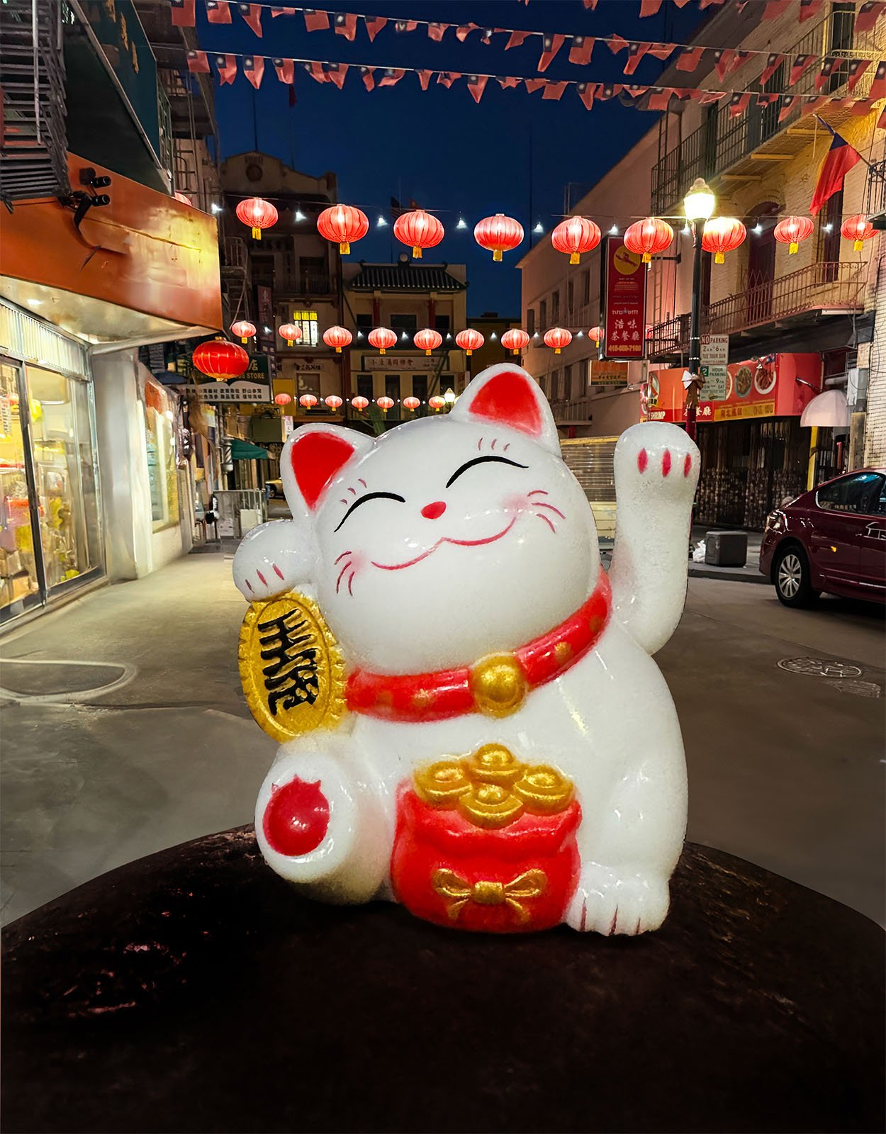 A white and red Maneki-neko (lure cat) figurine with raised paw and gold accents is prominent in the foreground.  In the background is a street decorated with red lanterns and warmly lit shops at dusk.