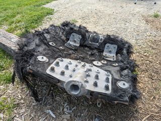 a large black piece of fiberglass covered in metal bolts and plates lies on the ground next to a path leading into a forest.  rolling mountains can be seen in the distance