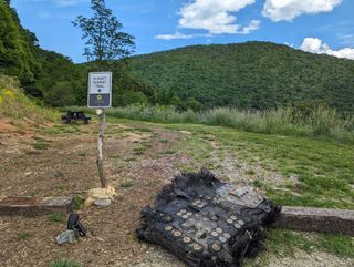 a large black piece of fiberglass covered in metal bolts and plates lies on the ground next to a path leading into a forest.  rolling mountains can be seen in the distance