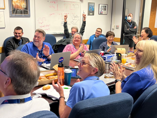 A bunch of people, many dressed in blue, cheer in a conference room.