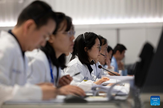 Six people in white outfits sitting in a mission control room, all looking in the same direction.