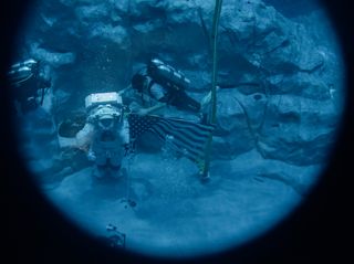 an astronaut in a spacesuit walks along the bottom of a pool next to a masked diver