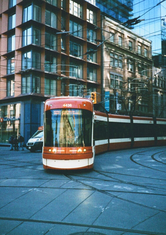 A red and white tram runs on rails on a city street corner, with modern and older buildings in the background.  A minibus is parked on the side of the street.  Reflections of buildings can be seen on the windows of the trams.