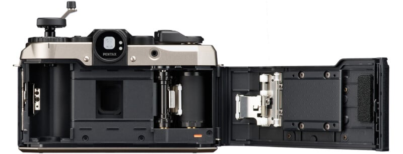 Open film camera showing interior with slots for film cans and reels.  The camera is labeled "Pentax" above the viewfinder.  The rear door is completely open, revealing the internal mechanisms and components.  The exterior of the camera is a combination of silver and black.