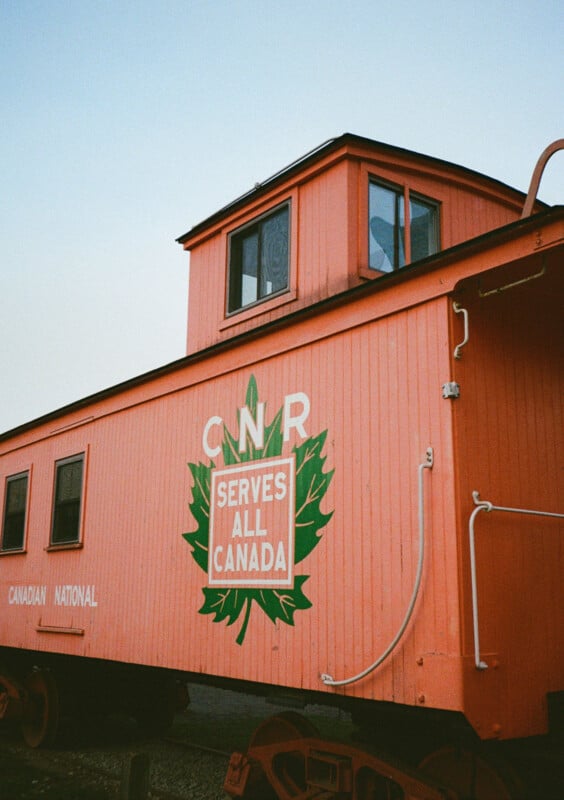 Canadian National Railway orange caboose with "CNR serves all of Canada" written in green graphics on the side of the leaf.  A caboose has a small cupola with windows on the roof and is located outside, with a clear sky in the background.