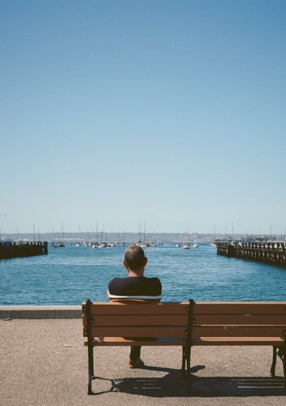 A man sits alone on a wooden bench facing a body of water with many boats anchored in the distance.  The sky is clear and blue, stretching over the calm water and stretching to the horizon.  Two pillars frame the stage on either side.