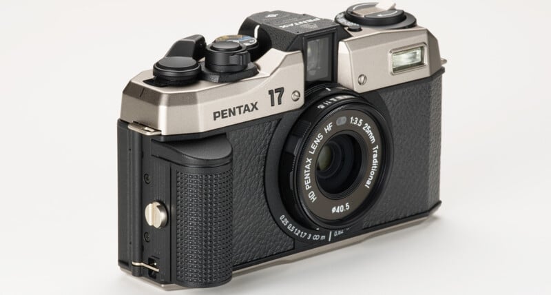 A silver and black Pentax 17 camera with a 12.5mm f/3.5 lens is shown on a white background.  The camera has a textured grip, multiple control dials on the top, viewfinder and flash on the front right above the lens.