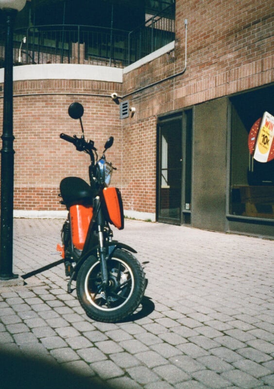 A red electric scooter is parked on a cobbled street, set against a background of red brick buildings.  To the right, a shop window displays a yellow sign advertising a 70% discount.  The area is brightly lit by the sun.