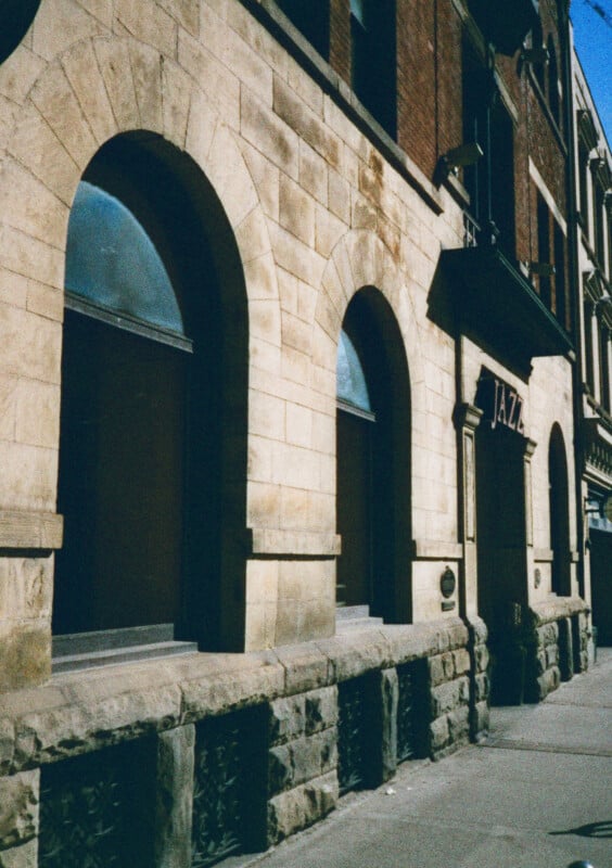 Street view of historic beige stone building with tall arched windows.  The building has an inscription "JAZZ" above the entrance.  The architectural style includes a mixture of stone and brick materials with decorative details.  Shadows fall on the sidewalk.