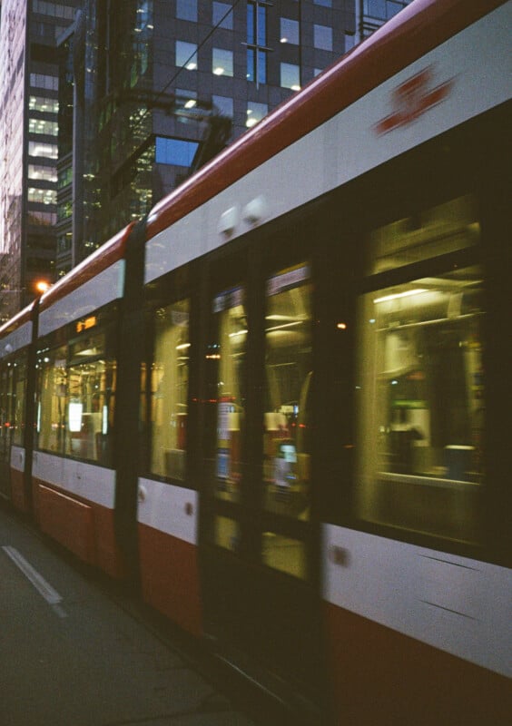 Blurred motion photo of a modern tram with red and white colors moving on a city street.  The background features tall buildings with lighted windows reflecting an urban evening scene.