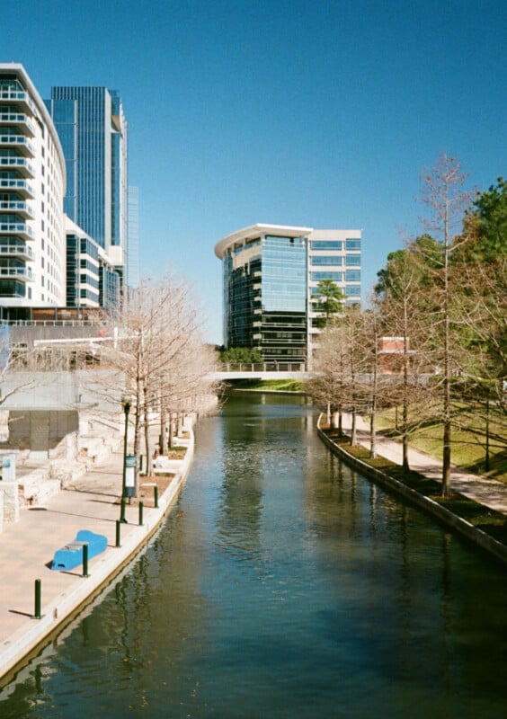 A quiet city canal lined with bare trees on both sides reflecting the clear blue sky.  Modern tall buildings lining the waterway with some greenery visible to the right.  A bridge in the distance connects the buildings over the canal.