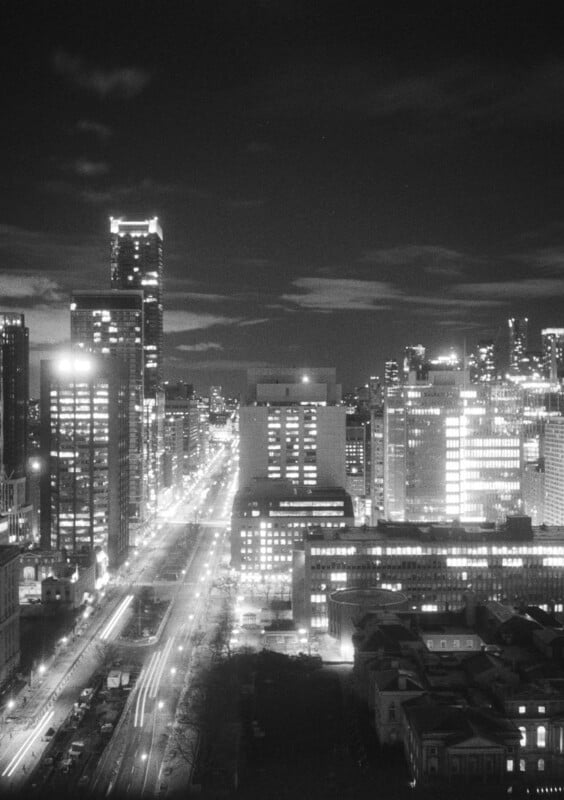 Aerial night view of a city showing illuminated skyscrapers and buildings lining both sides of a wide, well-lit street with the headlights of vehicles passing along it.  The sky is partly cloudy and the city lights create a glittering urban atmosphere.