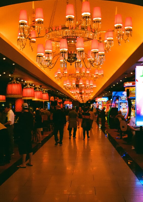 An interior scene of a busy casino includes people walking and talking among slot machines and gaming tables.  The space is illuminated by ornate chandeliers and warm lighting, creating a lively atmosphere.  The ceiling is decorated with red lampshades.