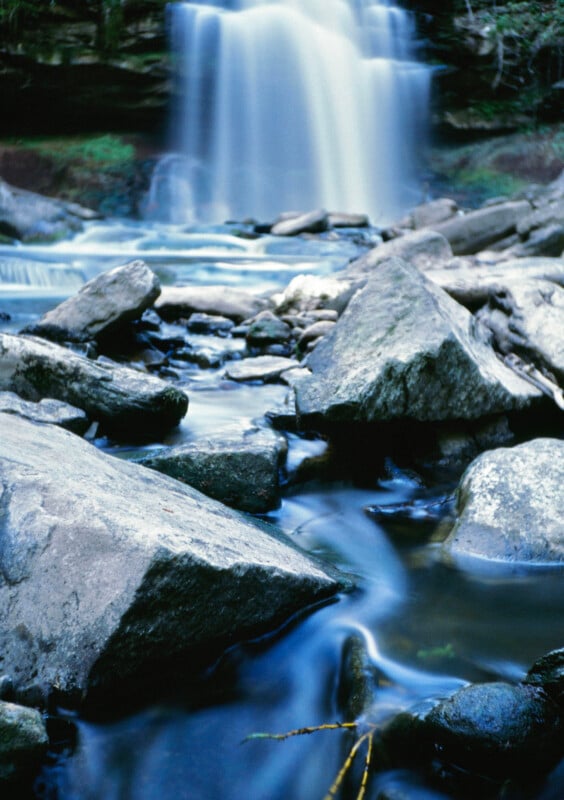 A tranquil waterfall cascades over a cliff into a rocky stream.  Large stones and boulders are scattered in the water, creating a calm and natural scene.  Lush greenery in the background enhances the peaceful atmosphere.