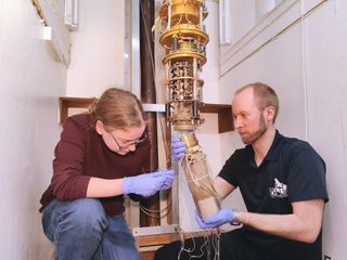 A white room containing two crouching people working on an elaborate golden machine