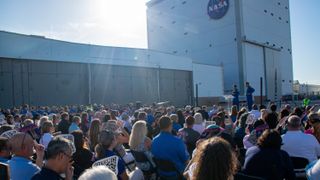 Two men in the distance stand in front of a tall building block in the sun.  It has a NASA logo on the top.  They address a large crowd, stretching out the rest of the frame.