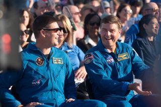 Two men in blue overalls laugh while sitting in a crowd.