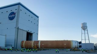 A large orange rocket booster stage lies horizontally on wheels outside a tall hangar and water tower.