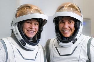 Two women in white spacesuits are seen from the chest up.  Their copper visors are turned up to show their faces padded in their helmets.  They are in a white room and they are smiling.