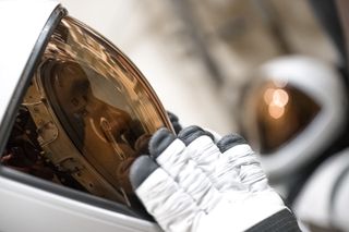 Close-up of an astronaut's helmet with a white shell on the left, extending to the middle of the image.  A copper-shaded visor extends across the darkened inner face.  A white glove with a black tip tests over the visor.