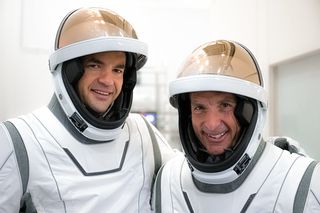 Two men in white spacesuits are visible from the chest up.  Their copper visors are turned up to show their faces padded in their helmets.  They are in a white room and they are smiling.